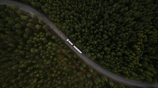 En lastbil syns från ovan köra på en slingrande väg genom granskog.